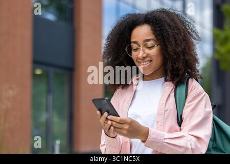 Eine lächelnde junge Frau mit Brille überprüft ihr Handy vor einem modernen Gebäude, trägt ein pinkfarbenes Hemd und einen Rucksack. Stockfoto