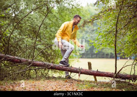 Mann mit hellgelber Regenjacke, der auf einen gefallenen Kiefernstamm am Waldrand tritt Stockfoto