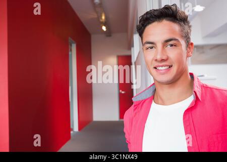 Mann lächelt, während er im modernen Flur des Büros steht, mit rotem Hemd und weißem T-Shirt, Kopierraum Stockfoto
