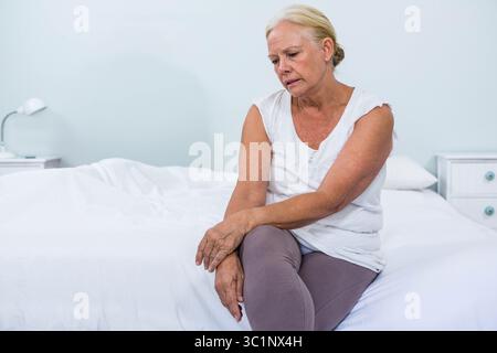 Ältere Frau, die auf der Bettkante im Schlafzimmer sitzt und das Handgelenk in der Nähe der Nachttischlampe und der Schubladen hält Stockfoto