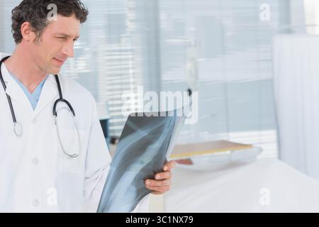 Arzt, der Röntgenfilm in der Brust neben dem Patientenbett im Krankenhauszimmer überprüft, mit Jalousien, die Licht streuen Stockfoto
