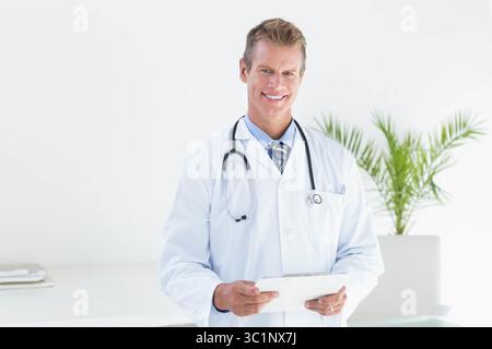 Reifer männlicher Arzt im Labormantel lächelnd, während er Tablette in der Klinik mit Stethoskop und Pflanze hält Stockfoto