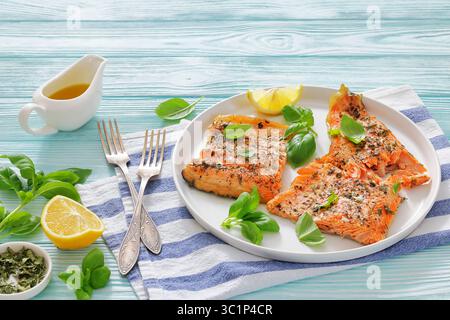 Gegrillte ganze Lachsfilets mit Haut auf einem weißen Teller mit frischem Basilikum, Zitronenscheiben, Gabeln auf Holztisch, horizontal von oben, Nahaufnahme, nicht A Stockfoto