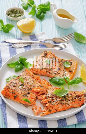 Gegrillte ganze Lachsfilets mit Haut auf einem Teller mit frischem Basilikum, Zitronenscheiben, Gabel und Gewürzen auf Holztisch, vertikale Ansicht von oben, schließen Stockfoto