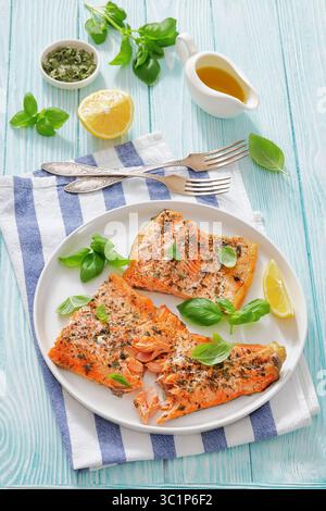 Gegrillte ganze Lachsfilets mit Haut auf einem Teller mit frischem Basilikum, Zitronenscheiben, Gabel und Gewürzen auf Holztisch, vertikale Ansicht von oben, keine Stockfoto