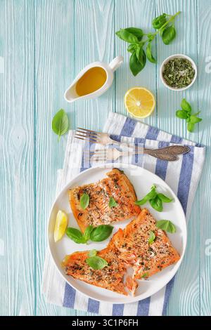 Gegrillte ganze Lachsfilets mit Haut auf einem Teller mit frischem Basilikum, Zitronenscheiben, Gabel und Gewürzen auf Holztisch, vertikale Draufsicht, freier Platz, Stockfoto