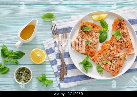 Gegrillte ganze Lachsfilets mit Haut auf einem Teller mit frischem Basilikum, Zitronenscheiben, Gabel und Gewürzen auf Holztisch, horizontale Ansicht von oben, fla Stockfoto