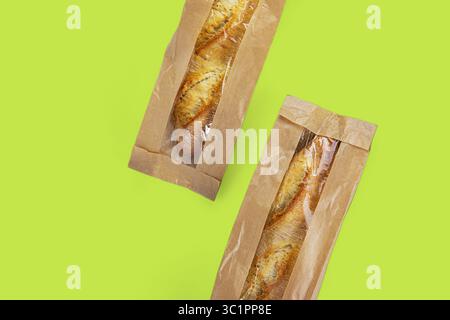 Zwei knusprige goldene Baguettes verpackt in umweltfreundlichen braunen Kraftpapiertüten mit transparenten Kunststofffenstern, isoliert auf einem leuchtend grünen Hintergrund. Stockfoto