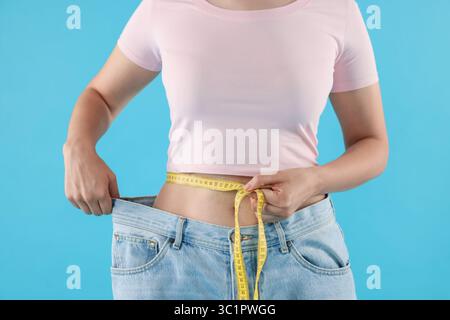 Frau mit Maßband, die alte Jeans nach Gewichtsverlust auf hellblauem Hintergrund trägt, Nahaufnahme Stockfoto