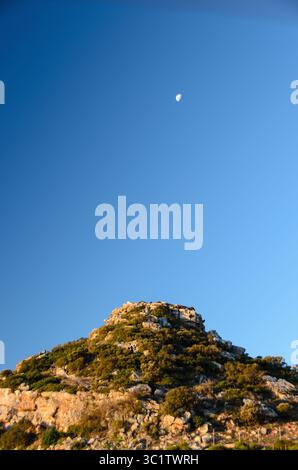 Ein sicheliger Mond scheint in einem klaren blauen Himmel über einem zerklüfteten, grünen Koppien und fängt eine ruhige Tageslandschaft ein. Stockfoto