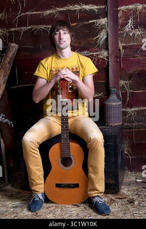 Ein junger Musiker sitzt selbstbewusst mit seiner Gitarre in einer rustikalen Scheune, in warmen Tönen. Stockfoto