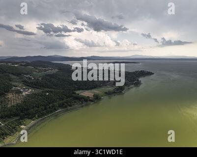 Aus der Vogelperspektive auf das ruhige olivgrüne Wasser des Sees küssen die grünen Küsten unter einem wilden Himmel, eine ruhige Landschaft entfaltet sich, Trasimeno See, Umbrien, Italien. Stockfoto