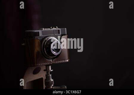 Eine Vintage-Kamera ist auf einem Stativ montiert und vor einem dunklen Hintergrund positioniert. Die Kamera ist in braunes Leder gewickelt und hat ein strukturiertes Objektiv. Stockfoto