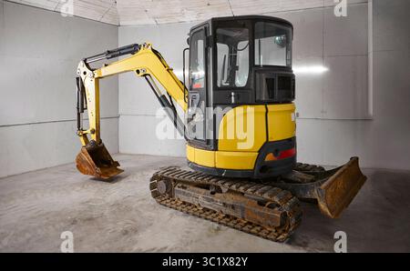 Ein gelber Minibagger befindet sich auf einem Betonboden in einem grauen, industriell aussehenden Raum. Der Bagger hat eine schwarze Kabine und einen hellgelben Arm und buc Stockfoto