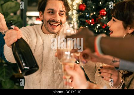 Ein glücklicher junger Mann mit einem fröhlichen Lachen toast mit einer Flasche Champagner an und feiert Weihnachten mit einer Gruppe von Freunden an einem lebhaften Teil Stockfoto