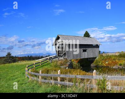 A.M. Foster Covered Bridge sitzt auf einer Wiese. Ein Holzzaun umgibt ihn. Die Brücke besteht aus verwittertem Holz, mit einem Fenster. Es erstreckt sich über einen kleinen Teich in Cabot, Stockfoto