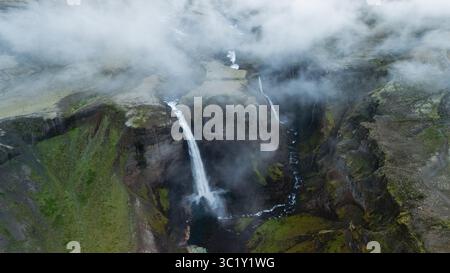Erleben Sie den atemberaubenden Blick auf den Haifoss-Wasserfall, der in einen tiefen Canyon eintaucht, umgeben von grünen Hängen und wirbelnden Wolken in der wilden Landschaft Islands. Ein wahres Naturwunder. Stockfoto