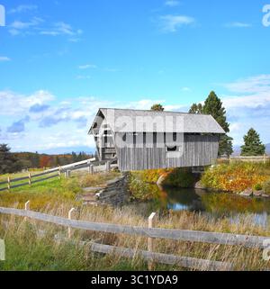 Die A. M. Foster Covered Bridge befindet sich in Cabot, Vermont. Holz und verwittert, überspannt die Brücke einen kleinen Teich. Herbstlaub steht im Hintergrund. Stockfoto