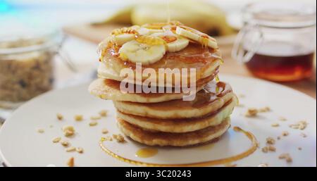 Pfannkuchenstapel auf Küchentisch mit Bananenscheiben und Sirup, mit Müsliglas Stockfoto