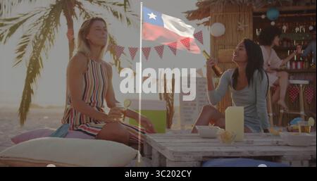 Lachende zwei Frauen, die Cocktails auf Palettentisch beim Sonnenuntergang am Strand halten, mit Tiki-Bar dahinter Stockfoto