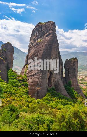 Eine dramatische vertikale Felsformation erhebt sich aus dichtem grünen Wald in Meteora, Griechenland, mit Bergen und verstreuten Wolken im Hintergrund auf einem warmen s Stockfoto