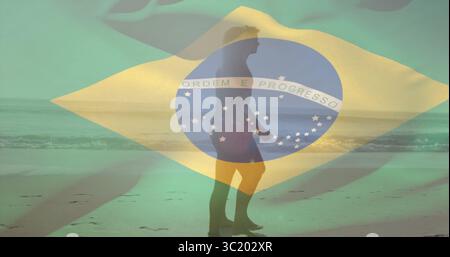 Walking Surfer in Boardshorts mit Surfbrett am Strand bei Ebbe, brasilianische Flagge überlagert Stockfoto