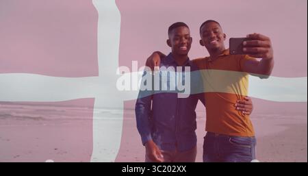 Selfie mit Smartphone machen, zwei Männer im orangefarbenen Polo am Sandstrand, mit dänischer Flagge Stockfoto