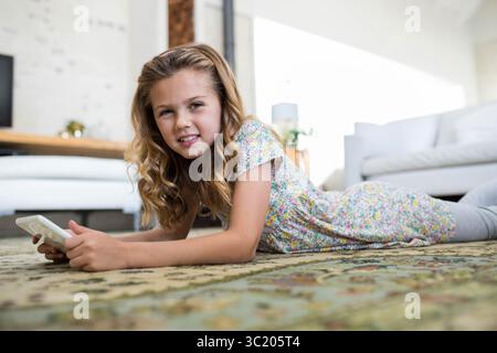 Weibliches Kind, das auf gemustertem Teppich im hellen Wohnzimmer liegt und ein weißes Smartphone hält Stockfoto
