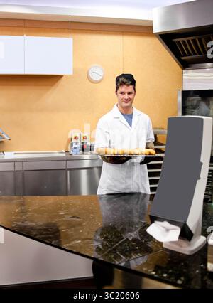 Erwachsener männlicher Bäcker mit Kochjacke, der hinter der Theke in der Bäckerei steht und ein Tablett mit Brötchen hält Stockfoto