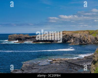 Zerklüftete Küstenklippen treffen unter hellem Himmel auf leuchtendes blaues Wasser in den malerischen Bridges of Ross, Irland. Stockfoto