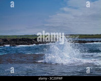 Mächtige Ozeanwellen brechen unter einem hellblauen Himmel an der felsigen Küste von Bridges of Ross, Irland, ab. Stockfoto