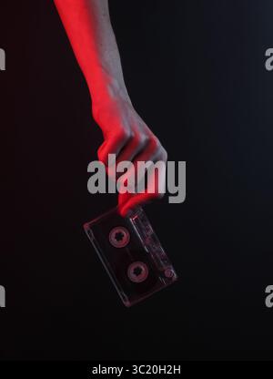 Weibliche Hand, die eine Retro-Audiokassette auf schwarzem Hintergrund mit rotem Neonlicht hält Stockfoto