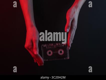 Weibliche Hände halten eine Retro-Audiokassette auf schwarzem Hintergrund mit rotem Neonlicht Stockfoto