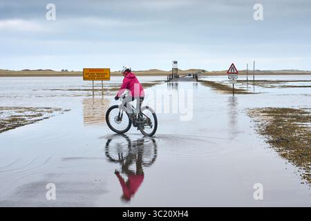 Frau mit Fahrrad, die ein Auto beobachtet, überquert den überfluteten Causeway nach Holy Island, Northumbria, England, Großbritannien Stockfoto