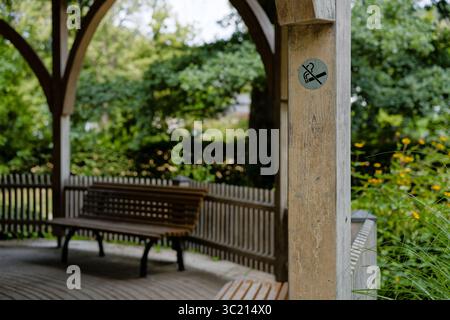 Bank in der Nähe eines Graduiertenturms in einem Park mit einem Rauchverbot auf einer Holzsäule, ruhige und entspannende Outdoor-Szene. Stockfoto