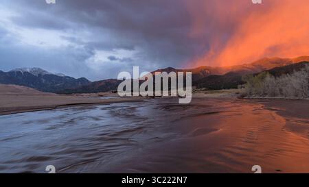 Sunset Medano Creek - Ein Sonnenstrahl beleuchtet nebelige Wolken, die über sanften Hügeln schweben und an einem stürmischen Frühlingsabend den Medano Creek rauschen. Stockfoto