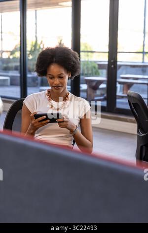 Afroamerikanerin hält Touchscreen-Gerät, während sie auf einem Mesh-Stuhl am Schreibtisch sitzt Stockfoto