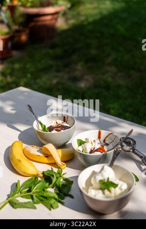 Eisschalen mit Schokoladensoße, frischer Minze und Bananen auf einem Holztisch in sonniger Umgebung, die ein erfrischendes Dessert schaffen Stockfoto