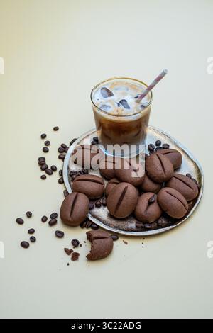 Eiskaffee mit Strohhalm steht neben Schokoladenkeksen in Form von Kaffeebohnen auf einer silbernen Platte und schafft eine einladende Dessertatmosphäre mit Wwa Stockfoto