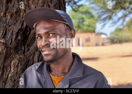 afrikanisches Dorf, junger erwachsener Mann mit Arbeitskleidung und Hut, Häuser im Hintergrund ländlich, im Schatten eines Baumes stehend Stockfoto