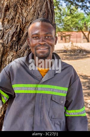 afrikanisches Dorf, junger erwachsener Mann in Arbeitskleidung, Häuser im ländlichen Hintergrund, im Schatten eines Baumes Stockfoto
