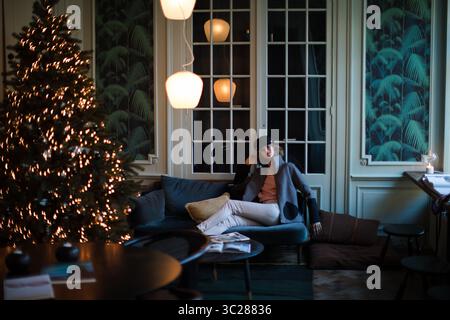 Modische Frau, die auf einem gemütlichen Sofa neben dem beleuchteten Weihnachtsbaum sitzt. In eleganter Umgebung. Das Modell ist entspannt, entspannt und selbstbewusst. Stockfoto