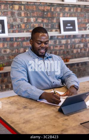 Afroamerikanischer Mann, der in Notizbuch am Holztisch im Arbeitsbereich schreibt und auf das Tablet am Stand blickt Stockfoto
