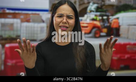 Frau spreizt ihre Hände und zeigt frustrierten Ausdruck in einer Bauszene, während der junge hispanic latin Influencer Emotionen in einem Outdoor-si ausdrückt Stockfoto
