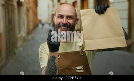 Ein lächelnder Mann hält Papiertüte auf der städtischen Straße mit Schürztätowierungen und Handschuhen, was auf einen modernen Lieferservice im Freien hindeutet. Stockfoto