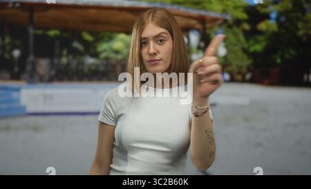Junge blonde Frau zeigt ihren Zeigefinger in einem sonnigen Park nach rechts; Anweisung Warnung Fokusbestimmung. Stockfoto