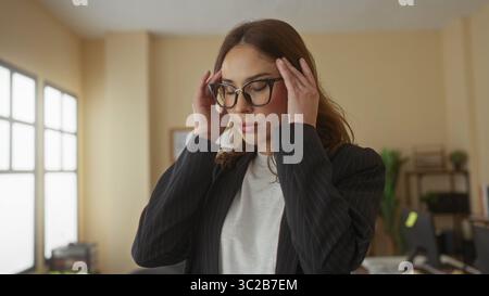 Frau, die Stress in einem Büro erlebt, den Kopf mit geschlossenen Augen hält, eine Brille und einen schwarzen Anzug in einem modernen Arbeitsplatz mit Keule trägt Stockfoto