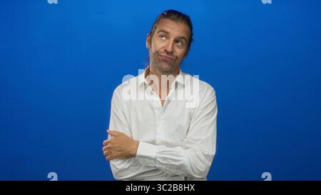 Ein blonder Mann mit langen Haaren, der ein weißes Hemd trägt, steht mit überkreuzten Armen in einem Atelier und drückt den Finger an die Lippen. Stockfoto