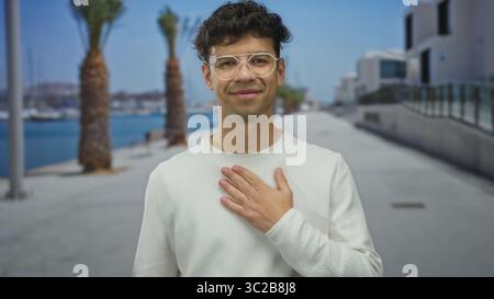 Mann Clutch Brust in weißem Pullover auf der Straße an der von Palmen gesäumten Strandpromenade am blauen Meer; Schmerzen. Stockfoto