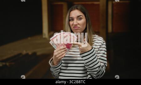 Frau mit isländischen Kronen-Banknoten auf einer Innenbaustelle mit einem verwirrten Ausdruck, der den unvollendeten Hintergrund kontrastiert. Stockfoto